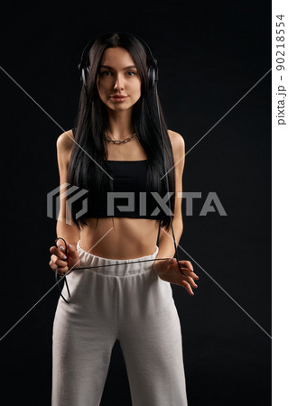 Young woman in headphones looking at the camera in studio. Portrait of brunette in black tank top standing straight, with earphones wire wrapped around hands, isolated on black. Concept of music fan. 90218554