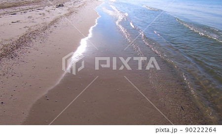 Waves of clean transparent water roll on the sandy beach of the sea shore. Person walking along the sea sandy beach. POV. Sun glare in the water. Seascape, waterscape background. Waves of clean transparent water roll on the sandy beach of the sea shore. Person walking along the sea sandy beach. POV. Sun glare in the water. Seascape, waterscape background. 90222264