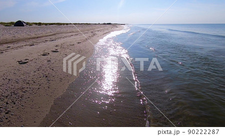 Waves of clean transparent water roll on the sandy beach of the sea shore. Person walking along the sea sandy beach. POV. Sun glare in the water. Seascape, waterscape background. 90222287