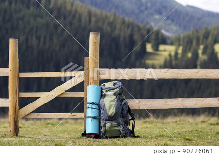 Big tourist backpack with attached carimate leaned at low wooden fence on lit by sun grassy valley on mountains covered with dense forest blurred background. Tourism, camping and traveling concept. Big tourist backpack with attached carimate leaned at low wooden fence on lit by sun grassy valley on mountains covered with dense forest blurred background. Tourism, camping and traveling concept. 90226061