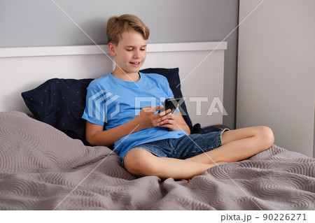 Teen boy uses smartphone for video chat while sitting on bed at home. Social life of a teenage. 90226271