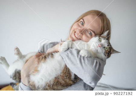 Portrait of young blonde happy 30s woman holding cute tabby cat with green eyes. Female hugging cat in hands 90227192