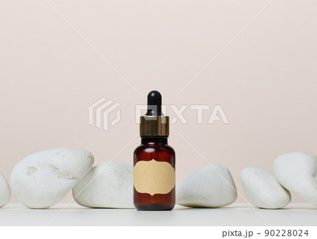 A brown glass bottle with a paper label and a pipette on a white table in the middle of the stones. A brown glass bottle with a paper label and a pipette on a white table in the middle of the stones. 90228024