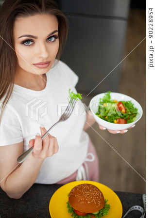 Beautiful young woman decides eating hamburger or fresh salad in kitchen. 90228954