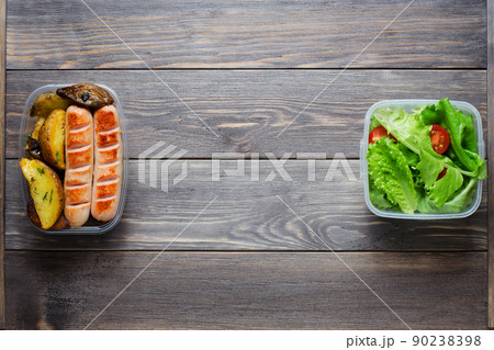Grilled sausages, potatoes, green lettuce with tomatoes in a plastic box. Unhealthy diet. Wooden background. Copy space 90238398