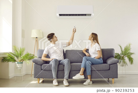 Young couple sitting on couch in living room at home switch on their air conditioner Young couple sitting on couch in living room at home switch on their air conditioner 90239253