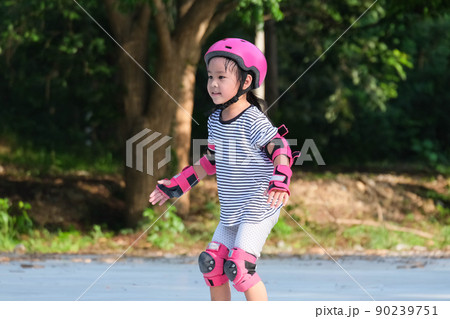 Cute Asian little girl in protective pads and safety helmet practicing roller skating in the park. Exciting outdoor activities for kids. 90239751