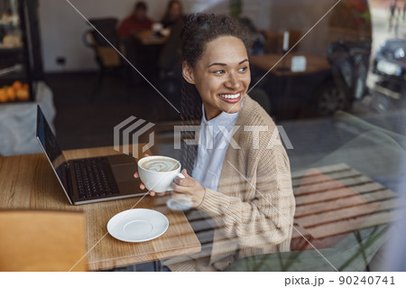 African cute woman works on laptop, smiles beautiful toothy smile, contemplates outside the window, enjoys coffee break African cute woman works on laptop, smiles beautiful toothy smile, contemplates outside the window, enjoys coffee break 90240741