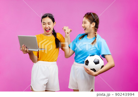 Two Cheerful Asian woman over isolated pink background holding a soccer ball and laptop, whatching football match, sport concept 90243994