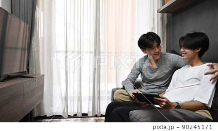 Happy young gay using digital tablet, watching movies on sofa together. Homosexual relationships and alternative love lifestyle concept 90245171