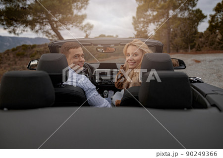 Happy couple of man and woman sitting in a convertible car and smiling while looking back Happy couple of man and woman sitting in a convertible car and smiling while looking back 90249366