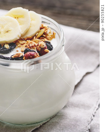 Natural homemade yogurt in a glass jar. Healthy food for breakfast with muesli. Jar with granola and banana slices on linen tablecloth on on wooden table. Natural homemade yogurt in a glass jar. Healthy food for breakfast with muesli. Jar with granola and banana slices on linen tablecloth on on wooden table. 90255821