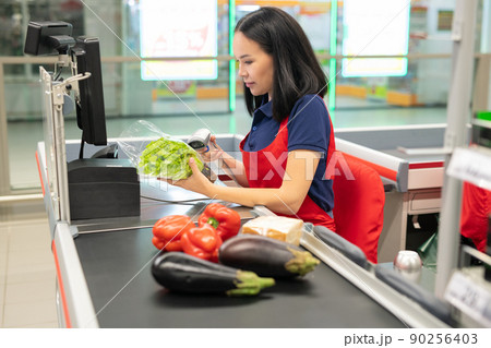 attractive Asian cashier wearing red apron selling fresh vegetables to customer attractive Asian cashier wearing red apron selling fresh vegetables to customer 90256403