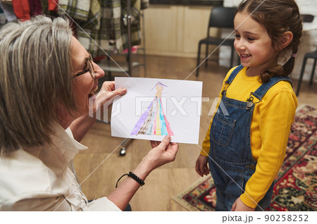 Pleasant elderly Caucasian woman tailor, caring and loving granny smiles admiring the clothes sketch of her adorable granddaughter while working in a fashion design studio and tailoring atelier 90258252