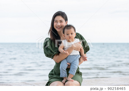 Happy cute baby boy with his mother on the beach. Summertime with baby is a quality time for Asian mommy.  Mother and baby on the beach and happy morning moment. Motherhood love and care for her child 90258465