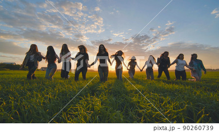 A group of girls walk towards the sun at sunset holding hands. 90260739