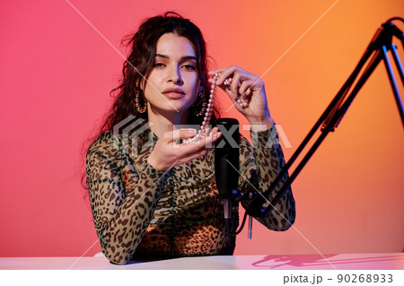 Modern young female blogger sitting in studio in pink and yellow neon light recording sounds for ASMR vlog using bead necklace 90268933