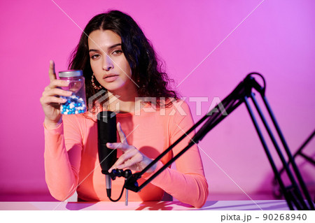 Young female blogger sitting in studio in pink neon light recording sounds shaking jar with pills for ASMR vlog 90268940