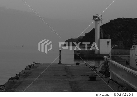 モノクローム　夕暮れの隠岐の島町漁港　桟橋に係留された小型漁船の船尾と防波堤上の白い灯台　静かな海面 90272253