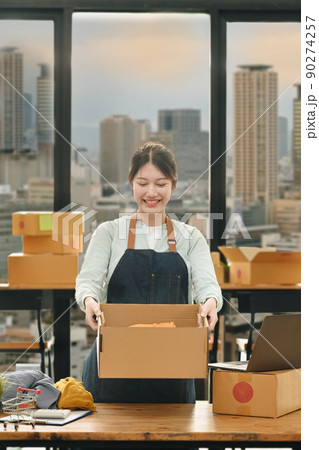 Smiling startup business owner holding cardboard and smiling to camera standing near large window at office with city buildings in background Smiling startup business owner holding cardboard and smiling to camera standing near large window at office with city buildings in background 90274257