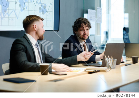 Young manager pointing at laptop and discussing with his partner presentation on monitor during conference at office 90278144