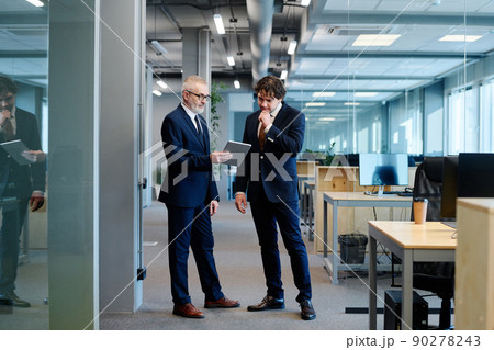 Mature businessman in suit showing something on digital tablet to his colleague while they standing at office Mature businessman in suit showing something on digital tablet to his colleague while they standing at office 90278243