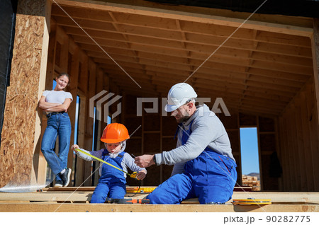 Father, mother and son building wooden frame house. Toddler boy helping his daddy, while woman looking for them on construction site. Guys wearing helmet and blue overalls. Carpentry, family concept. Father, mother and son building wooden frame house. Toddler boy helping his daddy, while woman looking for them on construction site. Guys wearing helmet and blue overalls. Carpentry, family concept. 90282775