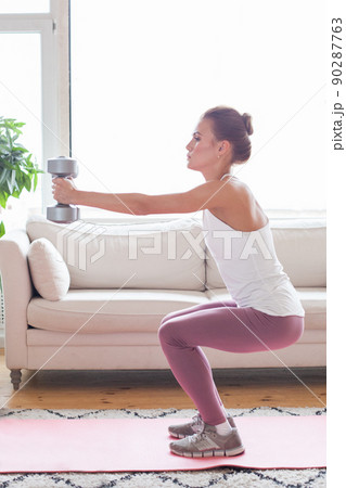 Young adult woman doing fitness exercises with gray dumbbells at her home in living room Young adult woman doing fitness exercises with gray dumbbells at her home in living room 90287763
