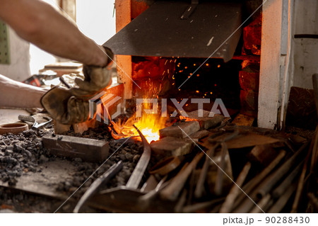 The blacksmith in production process of metal products, handmade in the forge. Craftsman heats metal in the fire. Metal industry, old profession. 90288430