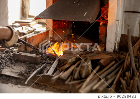 The blacksmith in production process of metal products, handmade in the forge. Craftsman heats metal in the fire. Metal industry, old profession. The blacksmith in production process of metal products, handmade in the forge. Craftsman heats metal in the fire. Metal industry, old profession. 90288433