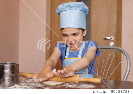 5 year old girl in blue dress and dressed as chef in kitchen laughs and rolling out dough 5 year old girl in blue dress and dressed as chef in kitchen laughs and rolling out dough 90288783