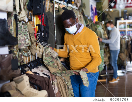 Male shopper in protective mask chooses camouflage uniform at army store Male shopper in protective mask chooses camouflage uniform at army store 90295793