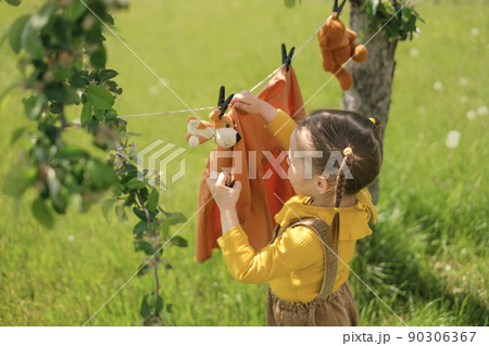 Girl with pigtails hangs washed wet clothes and soft plush toys on clothesline with clothespins under a tree in the garden. Small helper close-up. laundry of children's clothes and drying in fresh air 90306367