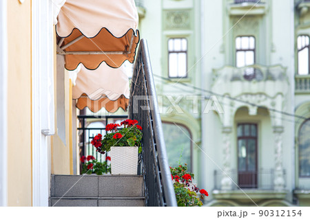 facade of building with an external staircase to balcony with black railing and decor on wall and decorative sun visors above windows and decorative flower pots with blooming red flower, nobody. facade of building with an external staircase to balcony with black railing and decor on wall and decorative sun visors above windows and decorative flower pots with blooming red flower, nobody. 90312154