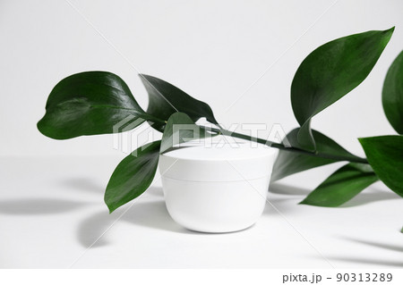 A mock up white jar of cream on a white gray background, on a white table with green leaves of a tropical plant, with hard shadows. Stylish look of the product, identity. 90313289