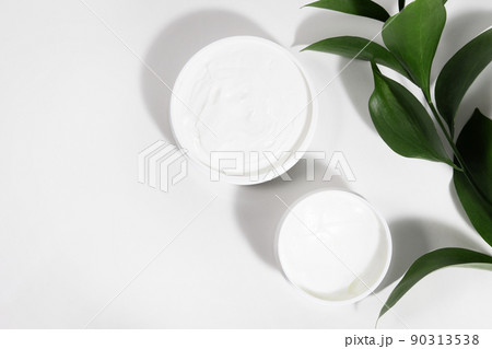A mock up white jar of cream on a white gray background, on a white table with green leaves of a tropical plant, with hard shadows. Stylish look of the product, identity. 90313538