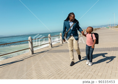 Happy african american father and son walking together on promenade against sky during sunny day 90335453