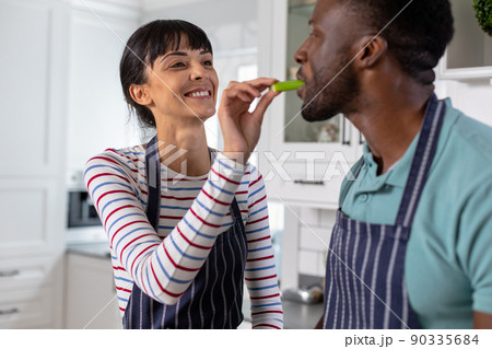 Smiling caucasian young woman feeding vegetable to african american boyfriend at home Smiling caucasian young woman feeding vegetable to african american boyfriend at home 90335684