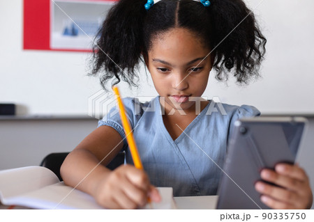 Biracial elementary schoolgirl using digital tablet while writing on book at desk in classroom Biracial elementary schoolgirl using digital tablet while writing on book at desk in classroom 90335750