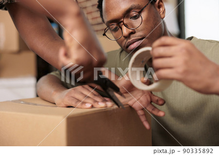 African american people sealing cardboard boxes with adhesive tape to move furniture in new real estate property. Packing things and shipping storage cargo, enjoying flat relocation. Close up. African american people sealing cardboard boxes with adhesive tape to move furniture in new real estate property. Packing things and shipping storage cargo, enjoying flat relocation. Close up. 90335892