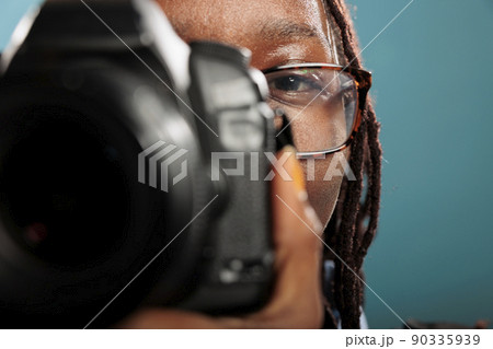 Close up of cheerful smiling professional photographer shooting photos using modern camera while standing on blue background. Young adult woman with photography hobby and DSLR device taking pictures. 90335939