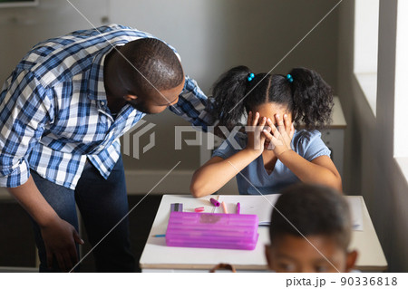 African american young male teacher consoling sad biracial elementary schoolgirl sitting at desk 90336818