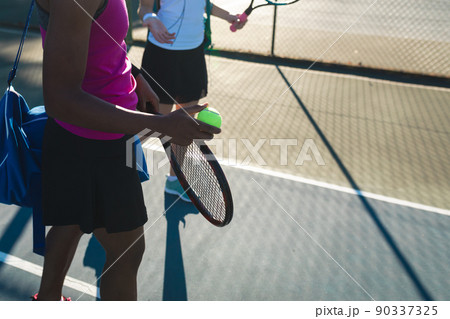 Biracial female tennis players standing at court on sunny day 90337325