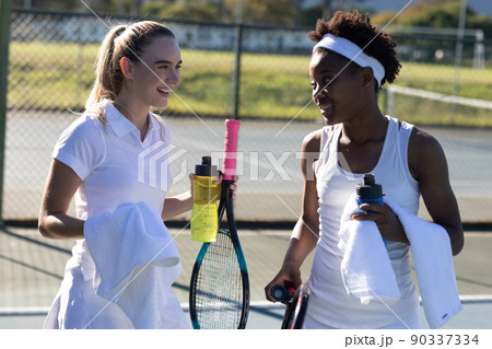 Smiling biracial female tennis players with bottles and towels talking during break at court Smiling biracial female tennis players with bottles and towels talking during break at court 90337334