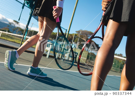 Low section of female multiracial tennis players walking rackets at court on sunny day Low section of female multiracial tennis players walking rackets at court on sunny day 90337344
