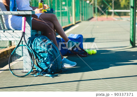 Low section of multiracial female players sitting on bench at tennis court during break on sunny day 90337371