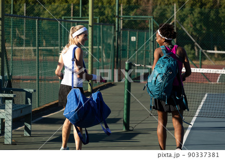 Smiling multiracial young female tennis players walking with bags at court on sunny day 90337381