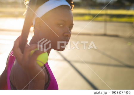 Portrait of young female african american player holding racket and ball at court Portrait of young female african american player holding racket and ball at court 90337406