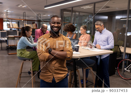 Portrait of african american businessman smiling while standing with arms crossed against colleagues Portrait of african american businessman smiling while standing with arms crossed against colleagues 90337453