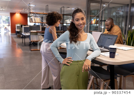 Portrait of smiling young hispanic businesswoman standing by desk against biracial colleagues 90337454